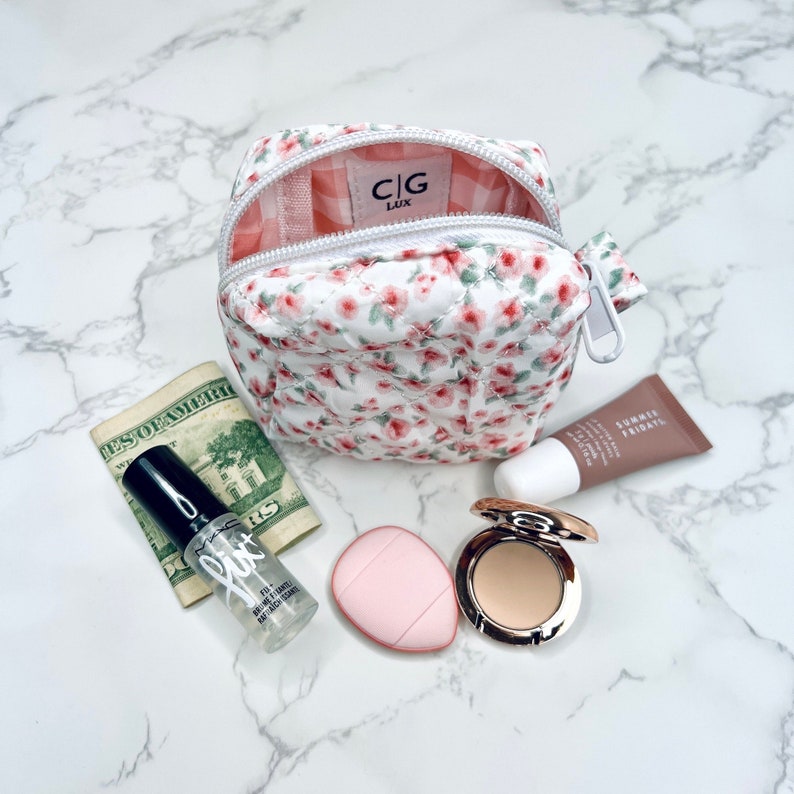 K&ouml;nnte beinhalten: Eine kleine wei&szlig;e, gesteppte Kosmetiktasche mit Blumenmuster, Rei&szlig;verschluss und rosa-wei&szlig;em Karomuster. Die Tasche ist offen und enth&auml;lt eine durchsichtige Flasche mit Make-up-Fixierspray, einen rosa Make-up-Schwamm, eine kompakte Foundation und eine Tube Lippenbalsam.