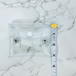 Puede incluir: Una peque&ntilde;a funda rectangular de pl&aacute;stico transparente con cierre de bot&oacute;n a presi&oacute;n. La funda mide aproximadamente 10 cm de altura. La funda est&aacute; vac&iacute;a y descansa sobre una superficie de m&aacute;rmol blanco. Una cinta m&eacute;trica est&aacute; al lado.