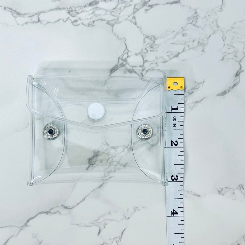 Puede incluir: Una peque&ntilde;a funda rectangular de pl&aacute;stico transparente con cierre de bot&oacute;n a presi&oacute;n. La funda mide aproximadamente 10 cm de altura. La funda est&aacute; vac&iacute;a y descansa sobre una superficie de m&aacute;rmol blanco. Una cinta m&eacute;trica est&aacute; al lado.