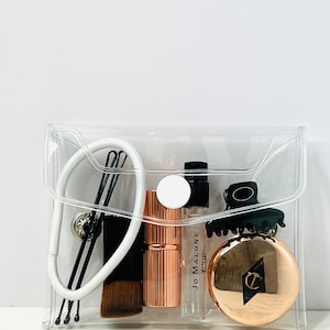 Puede incluir: Una bolsa de cosm&eacute;ticos transparente con varios productos de belleza. Incluye una goma para el pelo blanca, horquillas negras, una brocha de maquillaje, un labial oro rosa, un vial de perfume Jo Malone, una pinza para el pelo negra y un compacto oro rosa.