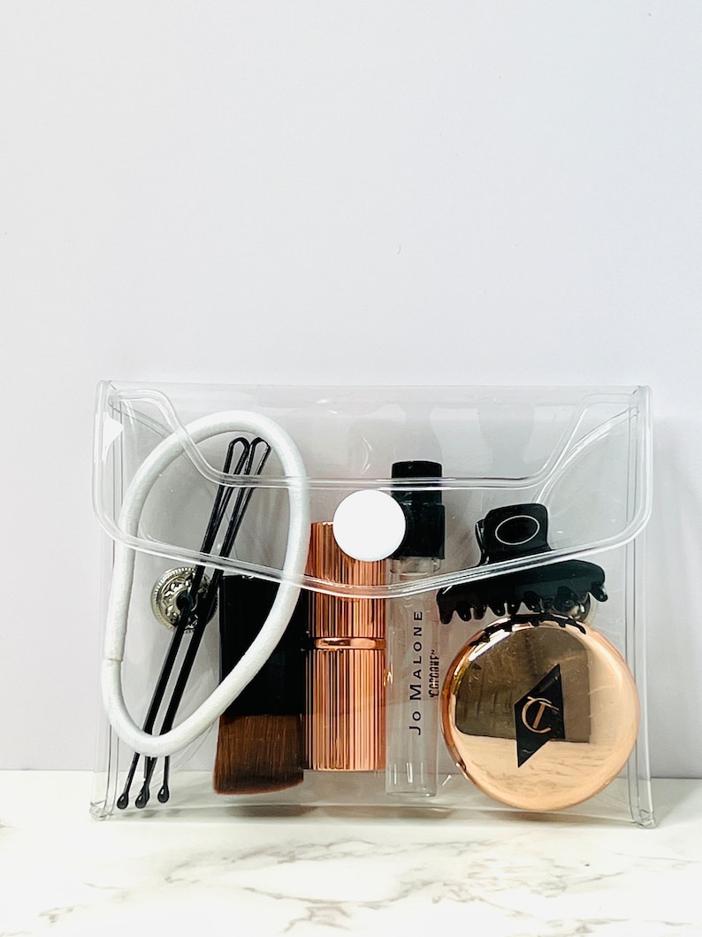 Puede incluir: Una bolsa de cosm&eacute;ticos transparente con varios productos de belleza. Incluye una goma para el pelo blanca, horquillas negras, una brocha de maquillaje, un labial oro rosa, un vial de perfume Jo Malone, una pinza para el pelo negra y un compacto oro rosa.