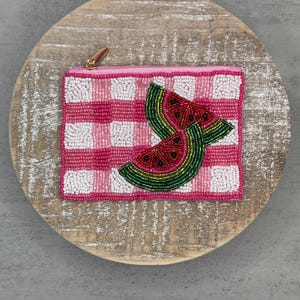 May include: A beaded pink and white gingham pattern pouch with a gold zipper. The pouch features a watermelon slice design with red, green, and gold beads. The pouch is on a round, light-colored wooden surface.