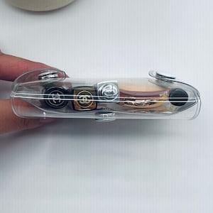Puede incluir: Un estuche cosm&eacute;tico rectangular transparente con cierres de bot&oacute;n plateados. El estuche contiene varias barras de labios y una polvera con logotipos de Chanel dorados y negros. El estuche est&aacute; sujeto por una mano sobre un fondo blanco.