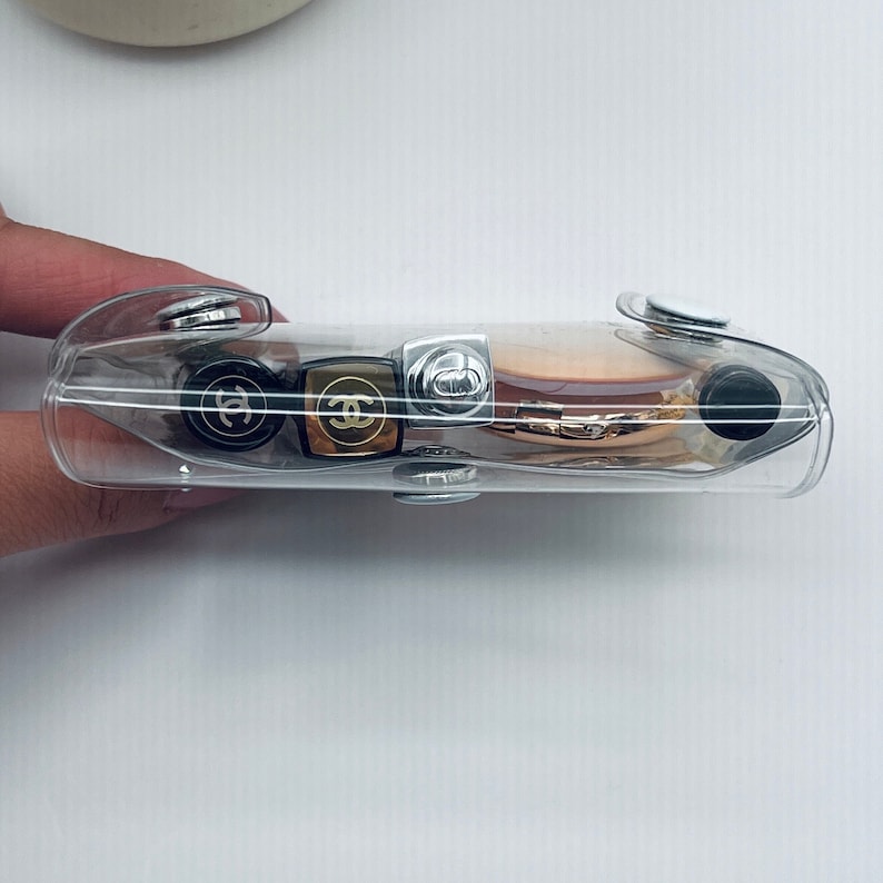 Puede incluir: Un estuche cosm&eacute;tico rectangular transparente con cierres de bot&oacute;n plateados. El estuche contiene varias barras de labios y una polvera con logotipos de Chanel dorados y negros. El estuche est&aacute; sujeto por una mano sobre un fondo blanco.