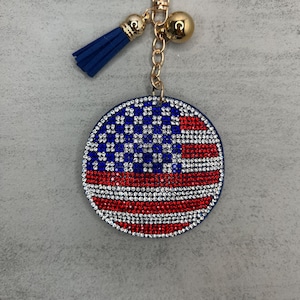 May include: A round keychain with a red, white, and blue American flag design made of rhinestones. The keychain has a gold chain and a blue tassel.