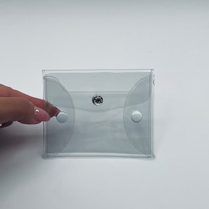 Puede incluir: Una funda de vinilo transparente con cierre de bot&oacute;n a presi&oacute;n. La funda tiene bordes redondeados y dos cierres de bot&oacute;n a presi&oacute;n blancos. Una mano con u&ntilde;as manicuradas sostiene la funda.