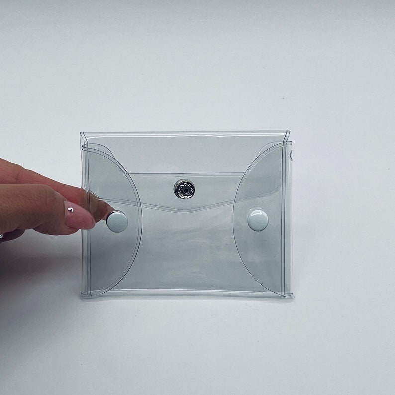 Puede incluir: Una funda de vinilo transparente con cierre de bot&oacute;n a presi&oacute;n. La funda tiene bordes redondeados y dos cierres de bot&oacute;n a presi&oacute;n blancos. Una mano con u&ntilde;as manicuradas sostiene la funda.