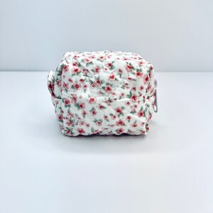 K&ouml;nnte beinhalten: Eine kleine wei&szlig;e gesteppte Tasche mit einem Blumenmuster aus rosa und gr&uuml;nen Bl&uuml;ten. Die Tasche hat einen Rei&szlig;verschluss.