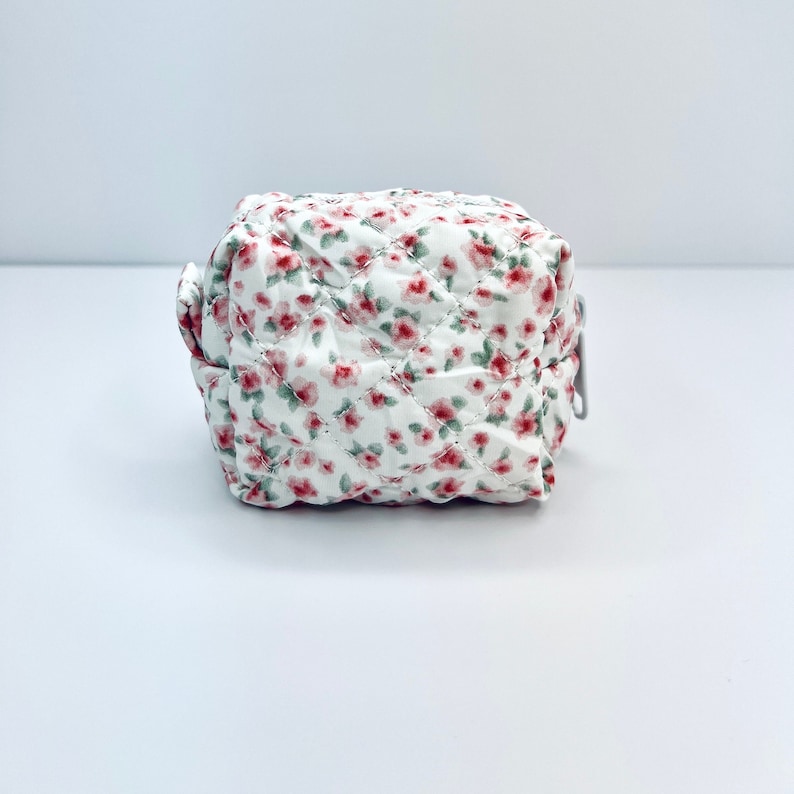 K&ouml;nnte beinhalten: Eine kleine wei&szlig;e gesteppte Tasche mit einem Blumenmuster aus rosa und gr&uuml;nen Bl&uuml;ten. Die Tasche hat einen Rei&szlig;verschluss.