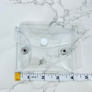 Puede incluir: Una peque&ntilde;a bolsa cuadrada transparente con cierre de bot&oacute;n a presi&oacute;n y dos broches plateados. La bolsa mide aproximadamente 10 cm de ancho. Una cinta m&eacute;trica amarilla y blanca se coloca junto a la bolsa.