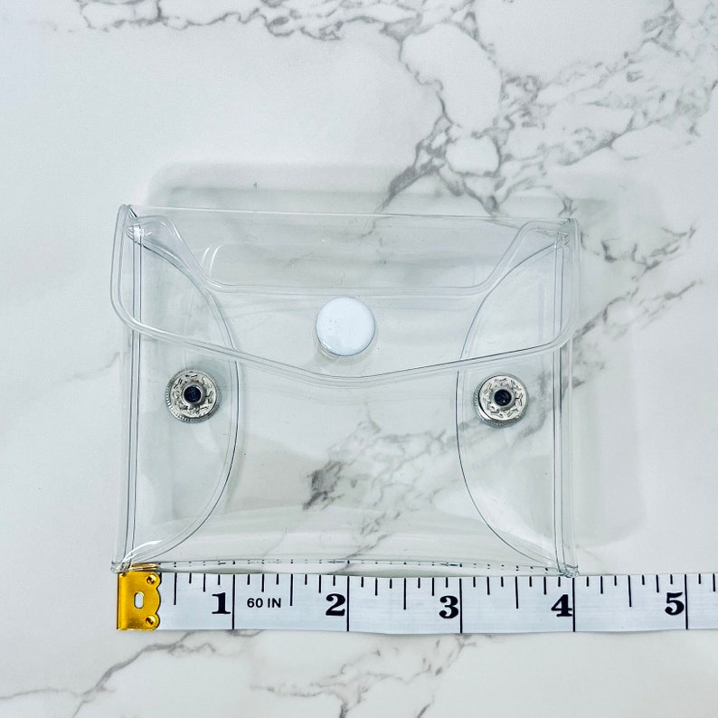 Puede incluir: Una peque&ntilde;a bolsa cuadrada transparente con cierre de bot&oacute;n a presi&oacute;n y dos broches plateados. La bolsa mide aproximadamente 10 cm de ancho. Una cinta m&eacute;trica amarilla y blanca se coloca junto a la bolsa.