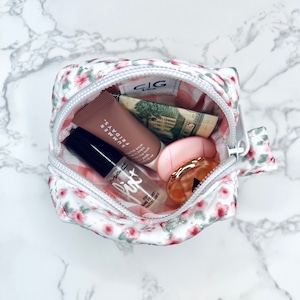 K&ouml;nnte beinhalten: Eine wei&szlig;e, mit Blumen bedruckte, gezippte Tasche mit einem rosa Lippenbalsam, einem klaren Lipgloss, einem kleinen Parf&uuml;mfl&auml;schchen und einem Dollar-Schein im Inneren.