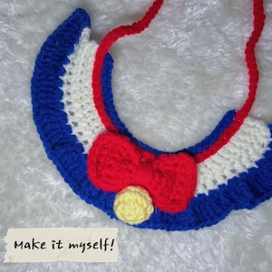 May include: A blue, white and red crocheted collar with a red bow and a yellow button. The collar is made of yarn and has a scalloped edge. The text "Make it myself!" is written on a piece of paper in the bottom left corner of the image.