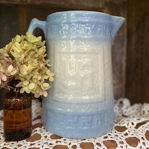 May include: A vintage blue and white ceramic pitcher with a handle and spout. The pitcher has a textured surface and decorative embossed details. A small brown glass bottle with dried flowers is next to the pitcher.