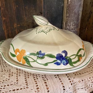 May include: Cream-colored ceramic butter dish with a lid featuring a leaf-shaped handle. The dish is decorated with orange and blue floral designs and green vines. The base has a scalloped edge and a green trim.