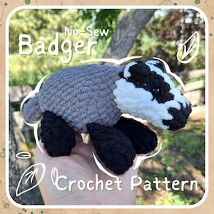 May include: No-Sew Badger Crochet Pattern. A crocheted badger plush toy with gray fur, black legs, and a white face. The badger is being held in a hand.