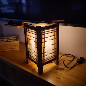 Könnte beinhalten: Eine Holzlaterne mit geometrischem Design. Die Laterne hat eine rechteckige Form mit einem gitterartigen Muster an den Seiten. Das Innere leuchtet mit einem sanften Licht. Eine passende Holzkiste ist im Hintergrund zu sehen.