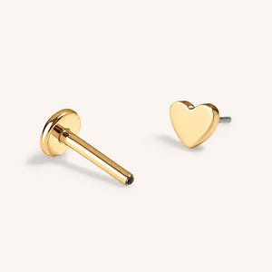 May include: A gold-toned flat back stud earring featuring a heart-shaped top and a smooth, cylindrical post. The piece is shown in two parts, displaying the heart-shaped front and the circular flat backing component separately on a plain background.