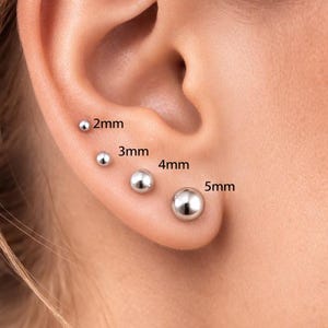 May include: A close-up of an ear with four silver ball stud earrings of varying sizes: 2mm, 3mm, 4mm, and 5mm. The earrings are displayed on the ear, showcasing their different dimensions and a polished finish.