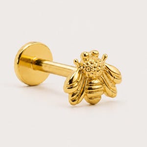 May include: A gold-toned labret stud earring featuring a bee-shaped charm. The bee charm has detailed wings and a textured body. The earring includes a flat circular back disc attached to a straight post, designed for body piercing applications.