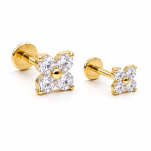 May include: Two gold-tone flat back labret studs featuring a four-petal flower design. Each flower is composed of four round, clear, faceted cubic zirconia stones surrounding a central gold-tone bead, set on a straight post with a circular base.