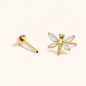 May include: A gold-toned dragonfly stud earring featuring four marquise-cut clear stones as wings and a beaded body design. The piece includes a flat-back post with a circular base, showcasing a polished metallic finish and clear faceted accents.