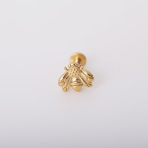 May include: A gold-toned bee-shaped stud earring featuring a detailed insect design with textured wings and a rounded body. The piece includes a flat circular backing post, showcasing a polished metallic finish throughout the entire jewelry item.