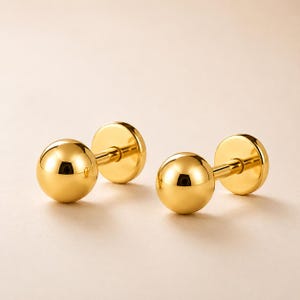 May include: A pair of polished gold-tone ball stud earrings featuring a smooth, spherical front design and a flat circular backing. The earrings have a reflective, metallic finish and a simple, clean construction with a post and secure back mechanism.