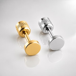 May include: Two barbell-style earrings, one in a polished gold tone and one in a polished silver tone. Each earring features a flat circular front, a smooth straight post, and a cylindrical back piece with a textured, diamond-patterned knurled edge.