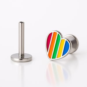 May include: A lip piercing stud with a heart-shaped design. The heart features a rainbow of colors: red, orange, yellow, green, and blue, with a silver-colored border. The stud is made of silver-colored metal.