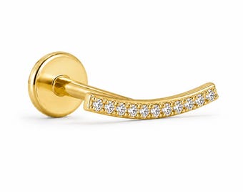 May include: A gold flat back labret stud featuring a curved bar design. The front of the curved bar is set with a row of ten small, clear, round-cut stones. The piece has a polished gold finish and a circular flat base at the end of the post.