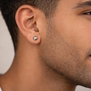 May include: A small, polished silver-tone ball stud earring worn in the earlobe. The earring features a smooth, spherical design with a reflective metallic finish, providing a simple and classic appearance for everyday wear in a single ear piercing.