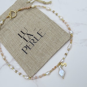 May include: A gold necklace featuring pearl and pink stone beads, a gold charm with the letter "S" and a large pearl pendant. The necklace is displayed on a beige fabric bag with the text "LU LA PERLE" printed on it.