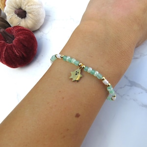 Op de afbeelding: Een kralenarmband met een gouden spookbedel wordt om een pols gedragen. De armband heeft lichtgroene en witte kralen met gouden accenten. Decoratieve fluwelen pompoenen in bruin-, crème- en bourgondische tinten zijn op de achtergrond.