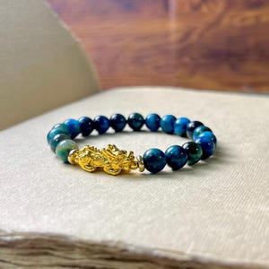 Może przedstawiać: Bransoletka z niebieskimi i zielonymi koralikami z tygrysiego oka, z pozłacanym urokiem. Tekst "Millionyearstone FENG SHUI JEWELRY" jest widoczny w tle. Bransoletka leży na białej powierzchni.
