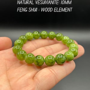 May include: A green vesuvianite bracelet with 10mm beads is held in a hand. The bracelet is made of round, polished beads. Text on the image reads: "NATURAL VESUVIANITE: 10MM FENG SHUI: WOOD ELEMENT."