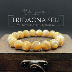 Può includere: Un braccialetto di perline gialle Tridacna sell, ciascuna di 10 mm di diametro, poggia su una scatola di legno. Le perline hanno un aspetto marmorizzato con toni bianchi e gialli. Il testo "TRIDACNA SELL" e "YELLOW TRIDACNA SELL BEADS 10MM" è visibile.