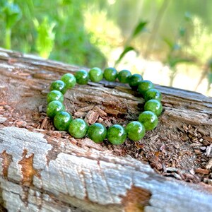 May include: A green beaded bracelet composed of round, polished beads. The bracelet is resting on a weathered, textured piece of wood, with a blurred green background.