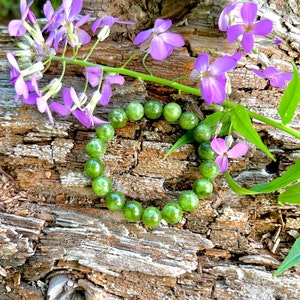 May include: A heart shape made of green gemstone beads rests on a weathered wood surface, with vibrant purple flowers and green leaves. The beads are round and polished, creating a contrast with the rough wood texture.