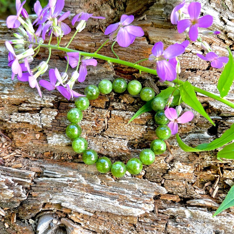 May include: A heart shape made of green gemstone beads rests on a weathered wood surface, with vibrant purple flowers and green leaves. The beads are round and polished, creating a contrast with the rough wood texture.