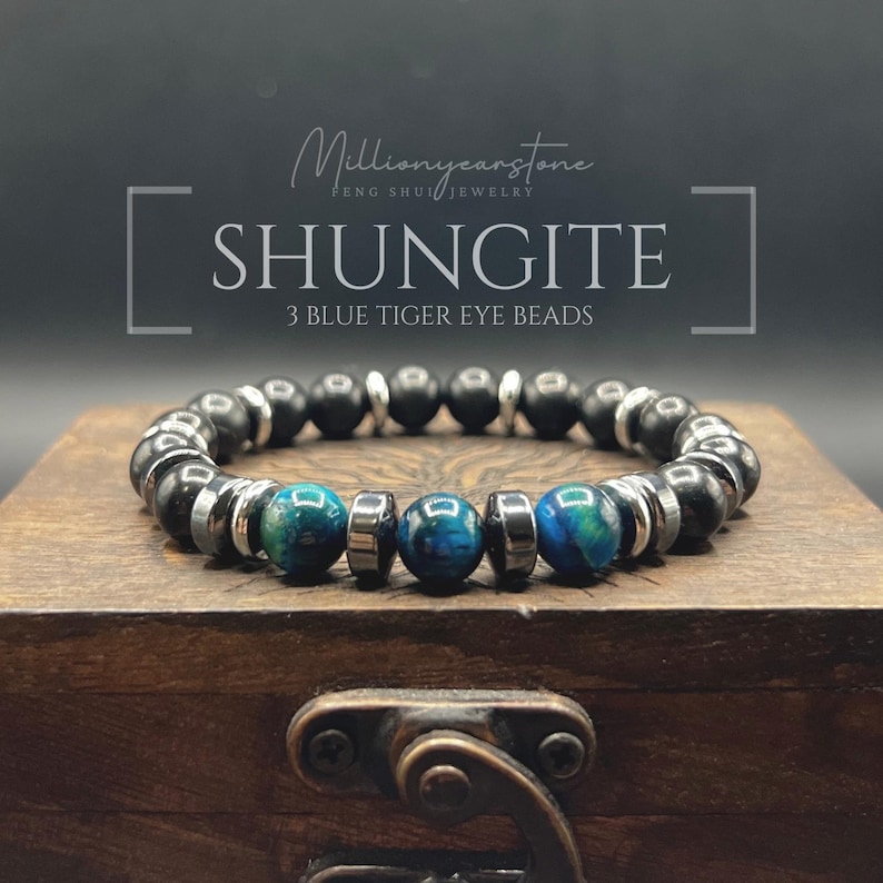 May include: Shungite bracelet with three blue tiger eye beads. The bracelet features black and silver beads. The text on the image reads "Shungite" and "3 Blue Tiger Eye Beads". The bracelet is displayed on a wooden box.
