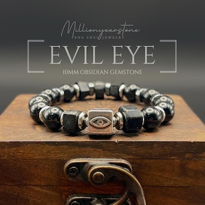 Puede incluir: Una pulsera de cuentas con esferas de obsidiana negra de 10 mm y cuentas negras facetadas, acentuada con espaciadores de metal plateado y un dije central cuadrado de color plateado grabado con un símbolo de ojo. El texto dice EVIL EYE.