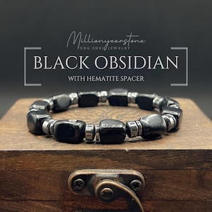 Könnte beinhalten: Ein Armband aus schwarzem Obsidian mit Hämatit-Abstandshaltern. Das Armband hat quadratische schwarze Obsidianperlen und kleine, runde, silberfarbene Hämatit-Abstandshalter. Das Armband wird auf einer Holzkiste präsentiert. Der Text auf dem Bild lautet "BLACK OBSIDIAN WITH HEMATITE SPACER."
