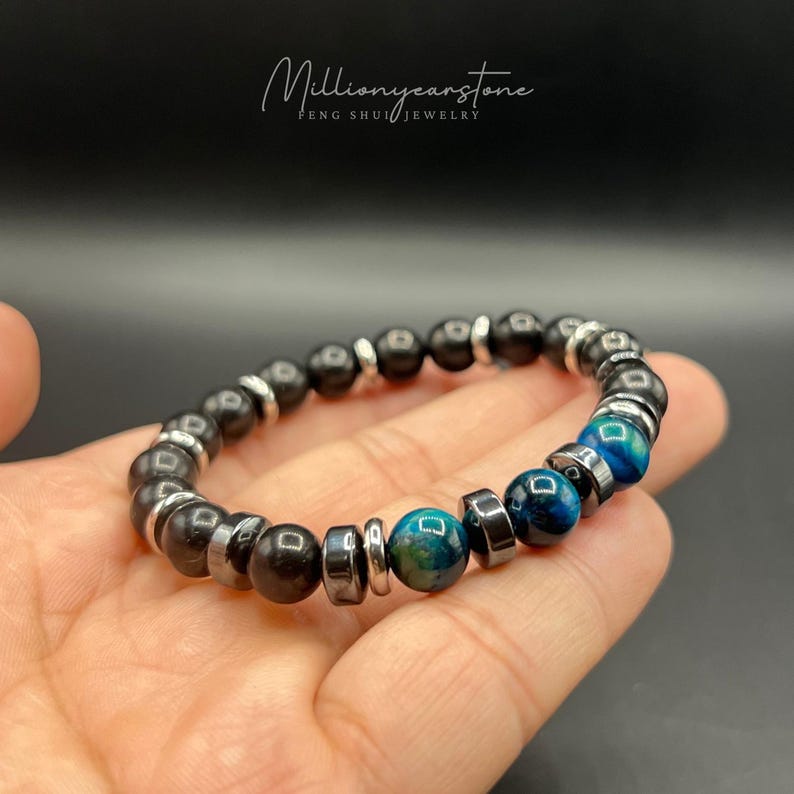 May include: A beaded bracelet featuring black and blue-green stones, interspersed with silver-colored metal beads. The bracelet is held in a hand, with the text "Millionyearstone FENG SHUI JEWELRY" in the upper left corner.