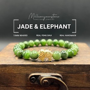 May include: A jade beaded bracelet with a gold elephant charm. The bracelet features 7.5mm beads and is labeled "Jade & Elephant", "Real Feng Shui", and "Real Handmade". The bracelet is displayed on a wooden box.