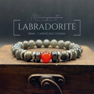 Puede incluir: Una pulsera de cuentas de labradorita con un dije de cornalina. La pulsera se exhibe sobre una superficie de madera. El texto en la imagen dice "LABRADORITE 8MM - CARNELIAN CHARM".