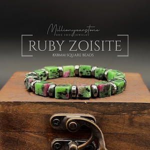 Op de afbeelding: Een armband met afwisselende vierkante robijn-zoisiet kralen en kleine metalen tussenstukjes. De tekst in de afbeelding luidt Millionyearstone FENG SHUI JEWELRY RUBY ZOISITE 8X8MM SQUARE BEADS. De kralen zijn groen met roze insluitingen.