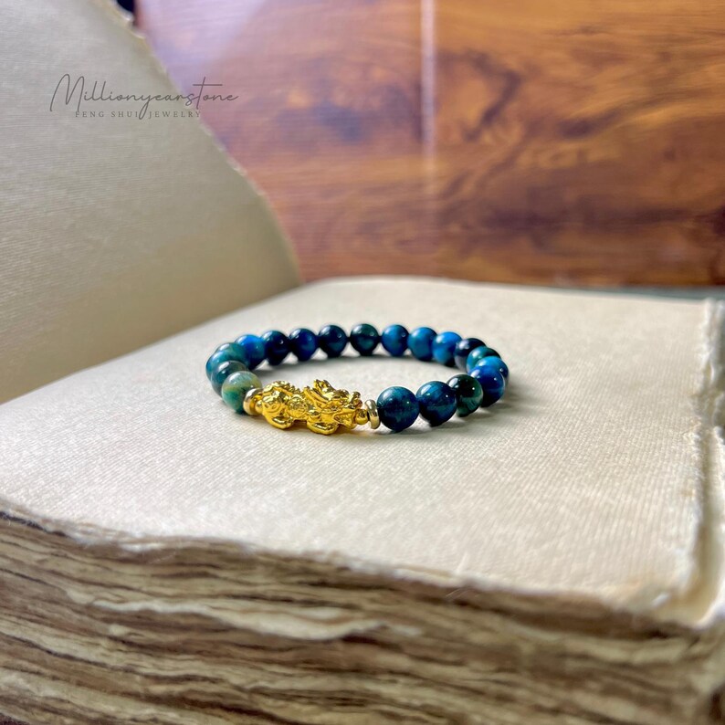 Może przedstawiać: Bransoletka z niebieskimi i zielonymi koralikami z tygrysiego oka, z pozłacanym urokiem. Tekst "Millionyearstone FENG SHUI JEWELRY" jest widoczny w tle. Bransoletka leży na białej powierzchni.