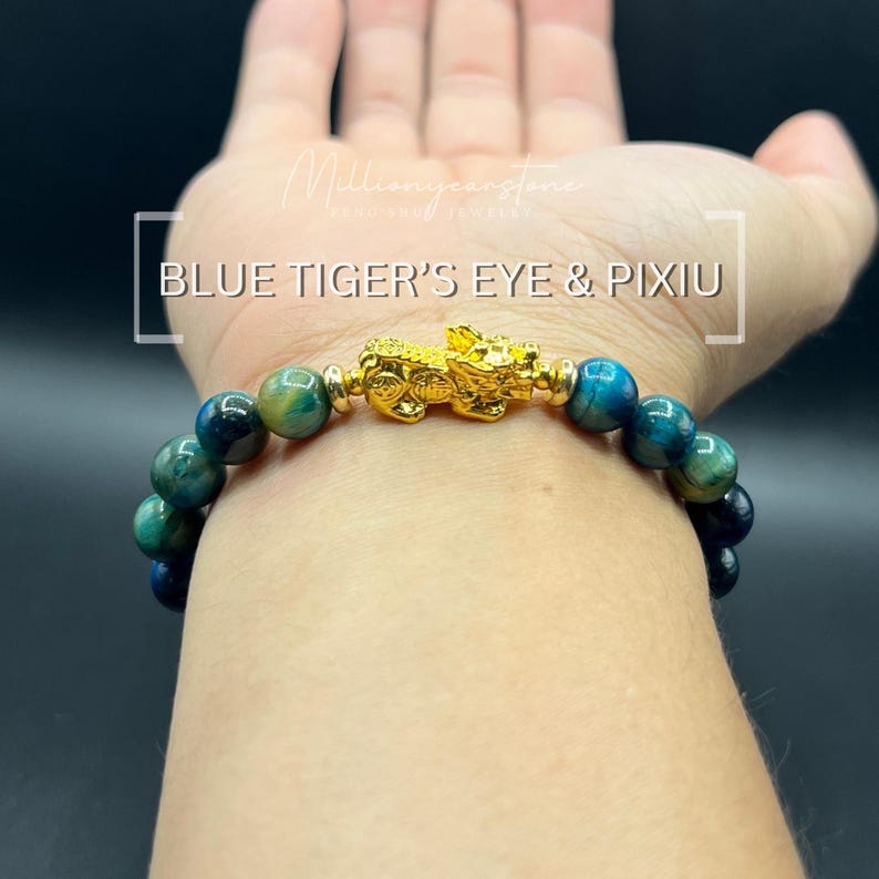 Może przedstawiać: Bransoletka z niebieskimi i zielonymi koralikami z tygrysiego oka oraz złotym urokiem Pixiu. Tekst "BLUE TIGER'S EYE & PIXIU" jest wyświetlany na biało. Bransoletka jest noszona na nadgarstku.