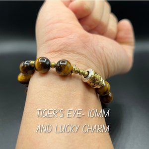 May include: A brown tiger's eye beaded bracelet with a gold and silver charm. The bracelet is 10mm in diameter. "TIGER'S EYE - 10MM AND LUCKY CHARM" is written on the image.